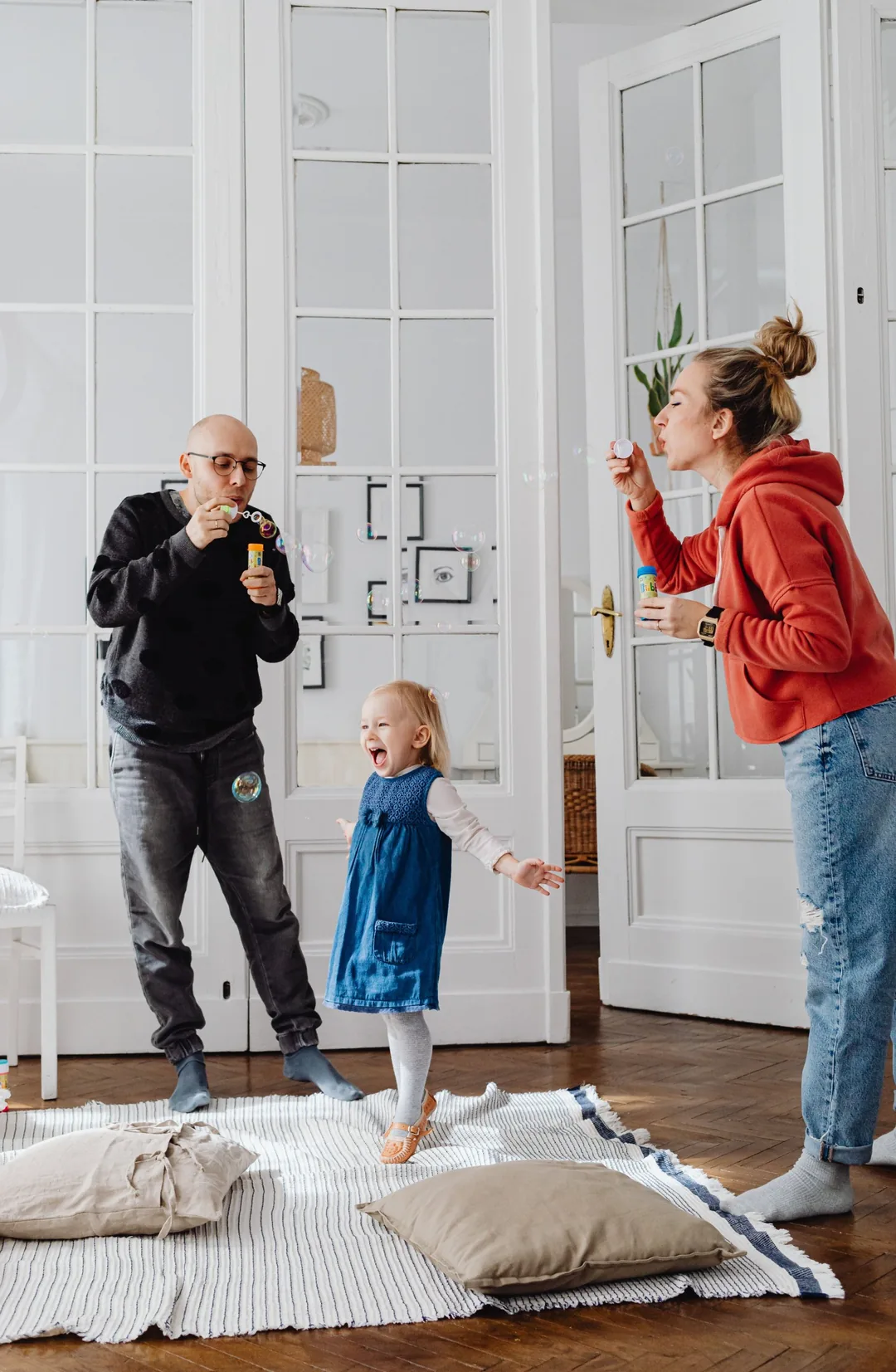 Des parents soufflent des bulles de savon autour de leur petite fille qui rit, dans un salon aux portes vitrées blanches et au parquet en point de Hongrie