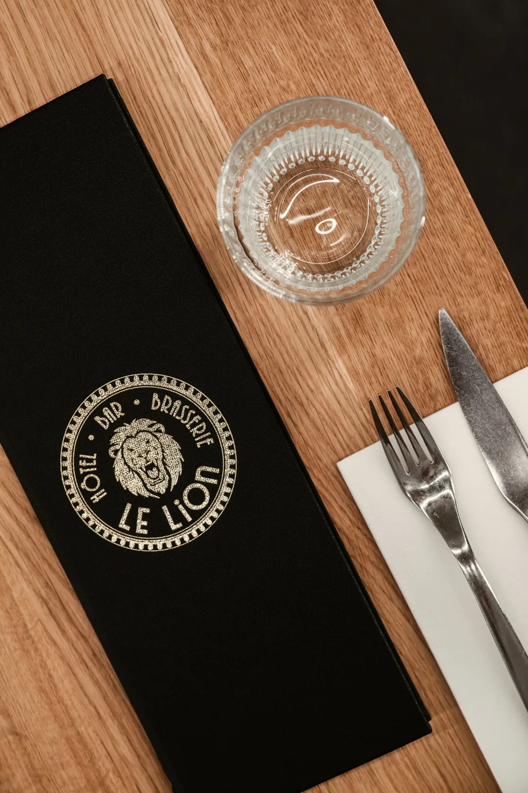 Porte-menu noir marqué du médaillon Le Lion, verre et couverts sur table en bois