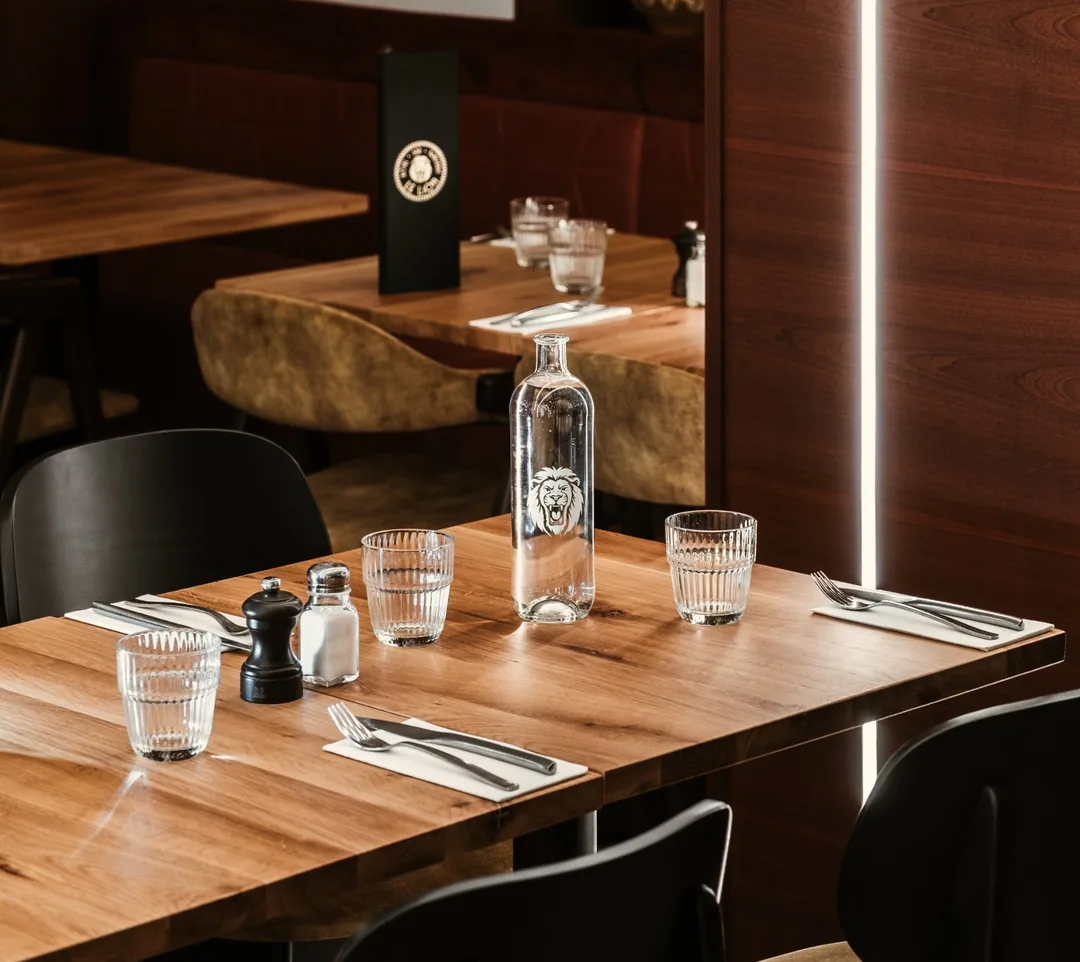 Salle du restaurant : tables en bois, carafe gravée au lion, verres